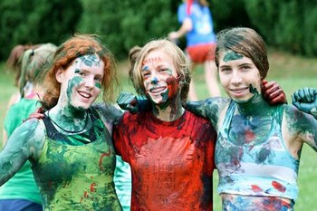 Na zdjęciu są trzy młode osoby, prawdopodobnie nastolatki, które mają na sobie ubrania pokryte kolorowymi farbami. Stoją blisko siebie i uśmiechają się do kamery, każda z nich z ręką na ramieniu sąsiada, co wyraża ducha drużynowości i wspólnoty. Twarze uczestników są również umalowane farbą, co sugeruje, że brali udział w zabawnej i kreatywnej aktywności, być może w ramach gry drużynowej lub festiwalu kolorów. Tło jest nieco rozmazane, ale widać zieloną przestrzeń, co wskazuje na to, że wydarzenie odbywa się na zewnątrz, w przyrodzie.